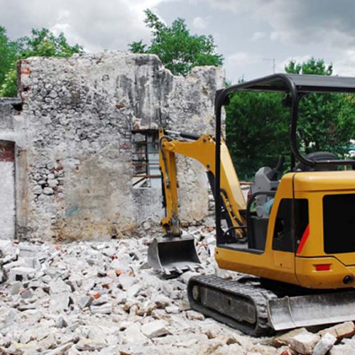 A compact excavator on a small domestic building site where a breezeblock building has just been demolished. The remains of an old Italian stone farmbuilding can be seen in the background